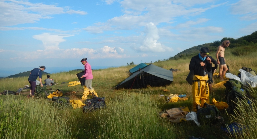 A couple of tarp shelters rest in a grassy meadow under blue skies. A few students stand nearby the shelters. 